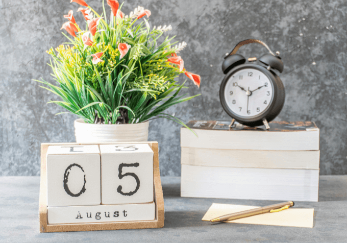 Wooden block calendar next to plant and a stack of books with a black alarm clock on top