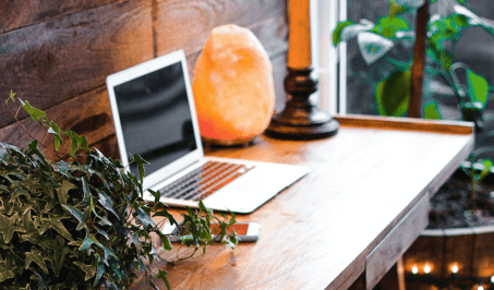 An open laptop on a desk, surrounded by plants and a glowing orange Himalayan salt lamp